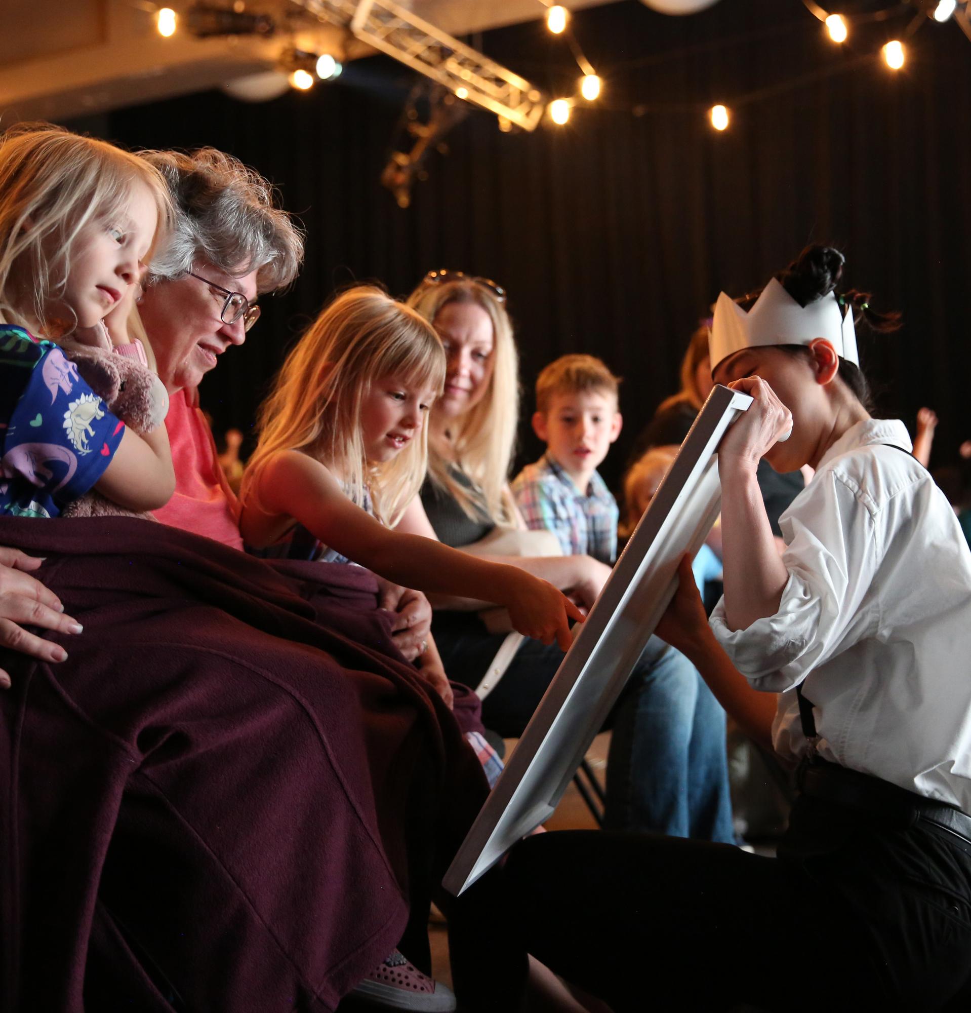 Children and families lean in as a performer in a paper crown shows them a drawing