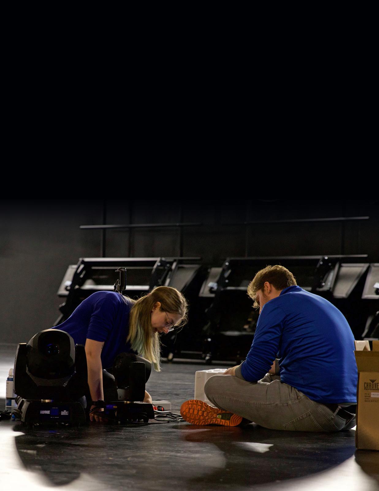 Two crew members set up stage lighting on an empty stage
