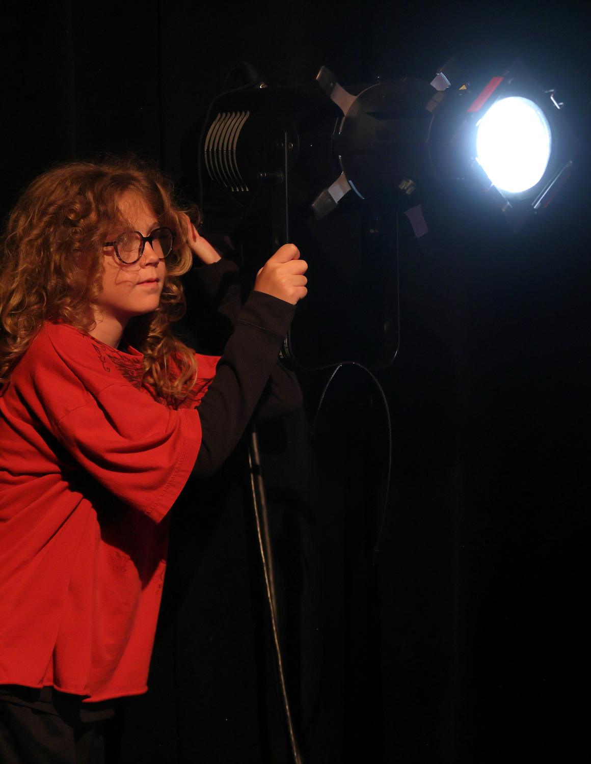 A young student in a red shirt adjusts a stage spotlight